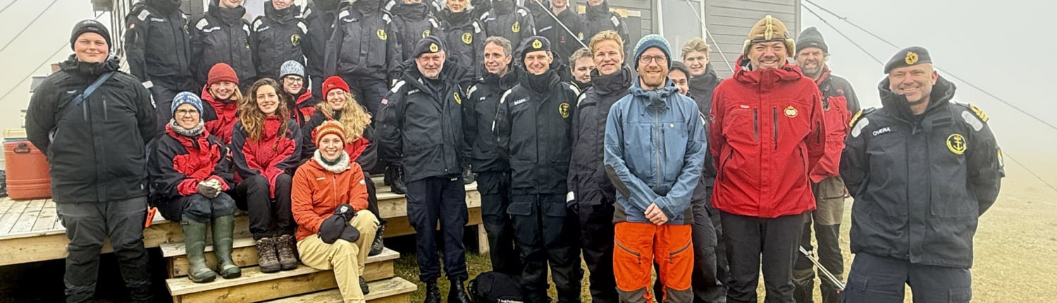 Gruppebilde av ca 30 personer som står/sitter foran en grå containerbygning med norsk flagg vaiende fra veggen
