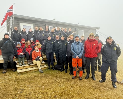 Gruppebilde av ca 30 personer som står/sitter foran en grå containerbygning med norsk flagg vaiende fra veggen